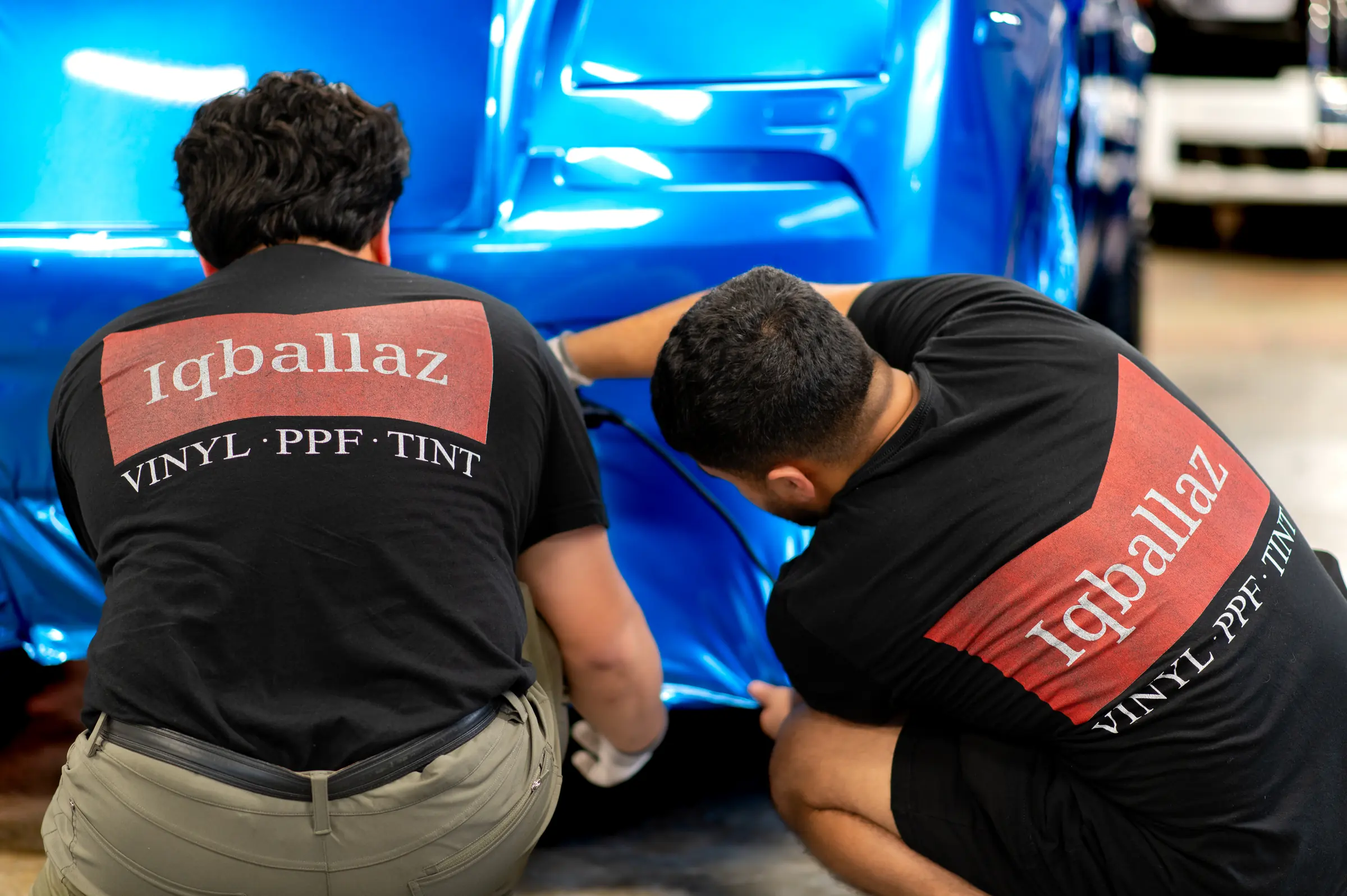Two Iqballaz installers laying blue vinyl over a front bumper